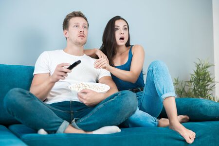 young couple beautiful brunette woman and handsome man watching movie on the sofa eating pop corn looking surprised cozy lifestyleの写真素材