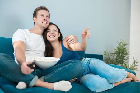 young couple beautiful brunette woman and handsome man watching movie on the sofa eating pop corn wife shows him exciting moment by pointing her fingerの写真素材