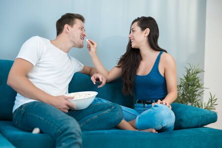 young couple beautiful brunette woman and handsome man watching movie on the sofa wife feeds husband with pop corn homestersの写真素材