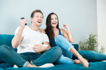 young couple beautiful brunette woman and handsome man cheer on their favorite team while watching football game eating popcorn at home cozy lifestyleの写真素材