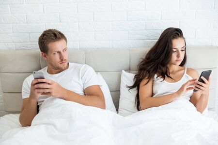 young couple handsome husband and his beautiful wife just woke up in bed checking their smartphones early in the morning man looks suspective social media addictionの写真素材