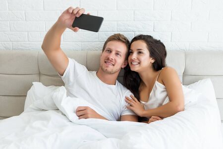 young happy couple handsome husband and his beautiful wife just woke up in bed shooting selfie on smartphone early in the morning social media addictionの写真素材