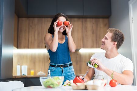 young funny couple cooking breakfast in the kitchen wife holds tomatoes near her eyes jokingly together smiling family traditionsの写真素材