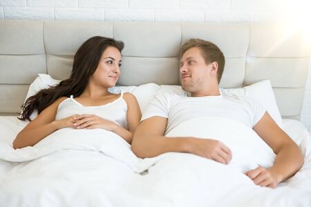 happy young couple handsome man and beautiful woman lying in bed in bright bedroom smiling to each other feeling love family idyllの写真素材