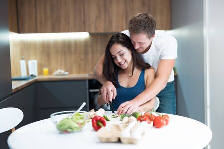 young couple cooking breakfast in the kitchen husband stands behind and hugs his beloved wife while they cut vegetables together family traditionsの写真素材