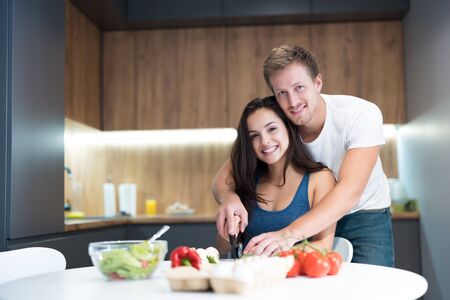 young couple cooking breakfast in the kitchen husband stands behind and hugs his beloved wife while they cut vegetables together smiling family traditionsの写真素材