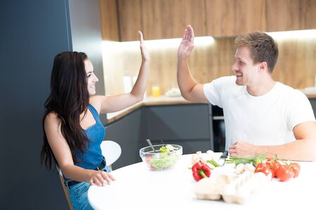 young couple handsome husband and his beautiful wife cooking breakfast in the kitchen together giving high five looking happy team workの写真素材