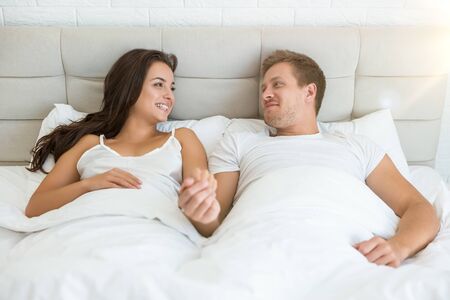 happy young couple handsome man and beautiful woman lying in bed in bright bedroom smiling to each other feeling love family morning idyllの写真素材
