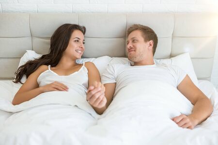 happy young couple handsome man and beautiful woman lying in bed in bright bedroom smiling to each other feeling love family morning idyll