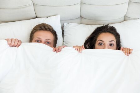 young couple handsome husband and his beautiful wife cuddling in bed in bright bedroom with blanket covering half of their faces family morning idyllの写真素材