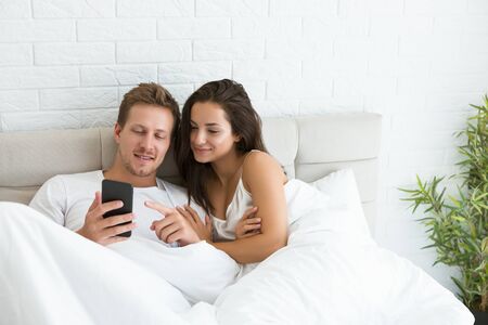 young couple handsome husband and his beautiful wife just woke up in bed watching photoes on smartphone early in the morning social media addictionの写真素材