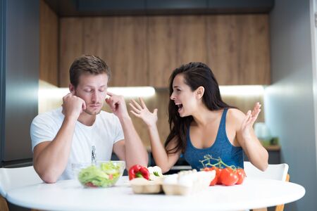 young couple having fight while cooking breakfast in the kitchen wife shouts loud at her husband he closes his ears with fingers looks annoyed family quarrelの写真素材