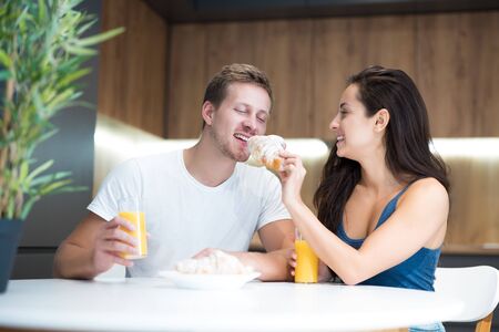 young lovely couple eating croissant and drinkink orange juice for breakfast in the kitchen woman feeds her husband start of great dayの写真素材