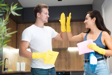 handsome young man giving high five to his beautiful wife wearing yellow gloves while doing cleaning in the kitchen together house choresの写真素材