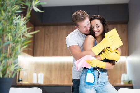 handsome young manhugging his beautiful wife wearing yellow gloves holding glass spray and rags...