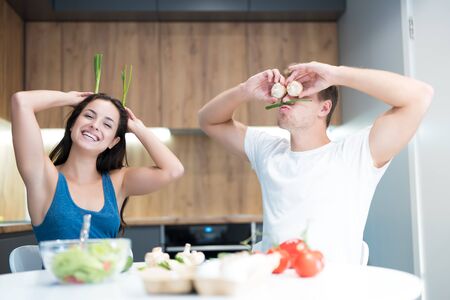 young funny couple cooking breakfast in the kitchen man closes his eyes with mushrooms while his beloved wife holds green onion like ears laughing togetherの写真素材