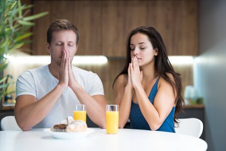 young couple saying prayer before start eating their breakfast in the kitchen family traditionsの写真素材