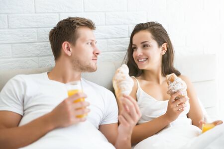 happy young couple handsome man and beautiful woman enjoying breakfast in bed eating croissant and drinking orange juice looking happy morning family idyllの写真素材
