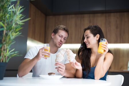 young lovely couple eating croissant and drinkink orange juice for breakfast in the kitchen playing with bagels start of great dayの写真素材