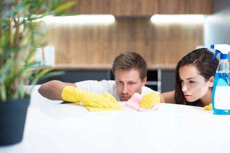 handsome young man and his beautiful wife wearing yellow gloves wipe off dust from kitchen table...