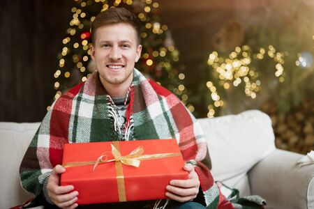 Young handsome man wearing warm sweater holding present sitting on the sofa in room decorated for...