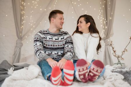 Young happy couple beautiful woman and handsome man both wearing warm sweaters and festive socks sitting in bedroom decorated for celebrating new year christmas tendernessの写真素材