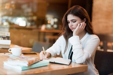 young beautiful woman in white stylish jacket working on project outside office writing in planner during coffee break in cafe looking tired modern businesswoman multitaskingの写真素材