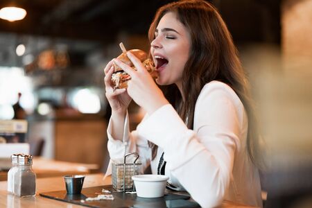 young beautiful woman in white stylish blouse looks hungry biting fresh meat burger during lunch in trendy cafe eating outsideの写真素材