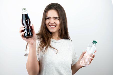 young woman with bottle of soda drink in one hand and bottle of water in another biting her lipon isolated white background dietology and nutritionの写真素材