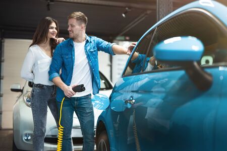 young couple handsome man and beautiful woman checking new vehicle standing near open gasoline hatch in car dealership center, looking happyの写真素材