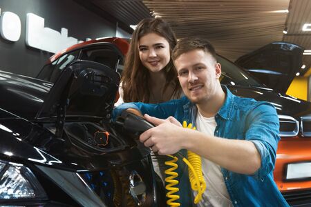 young happy family couple handsome husband shows his beautiful brunette wife how to use charging cable for electric car , looking excited, eco friendly conceptの写真素材