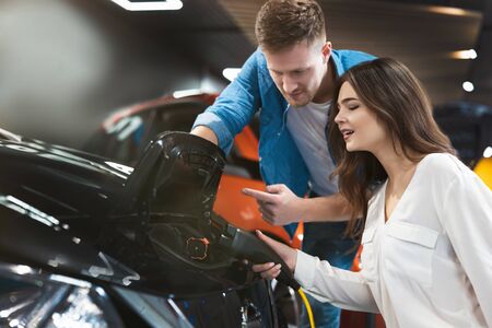 handsome man teaches beautiful brunette woman how to use charging cable for electric car sitting near open bonnet , looking happy and excited, eco friendly conceptの写真素材