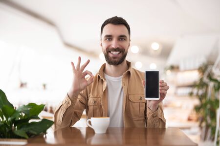 Young handsome satisfied man drinks coffee in the cafe out for lunch demonstrating his smartphone, showing like sign , communication and technology conceptの写真素材