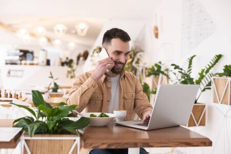 Young handsome bearded man working in his laptop talking on smartphone in his hand during lunch break at cafe, looking busy, multitasking conceptの写真素材