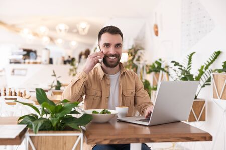 Young handsome bearded smiling man working in laptop talks on smartphone in his hand during lunch break at cafe, looking busy and satisfied, multitasking conceptの写真素材