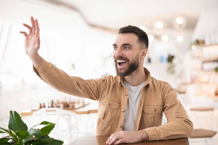 Young handsome bearded man waving greeting his friend while sitting in the cafe , body language conceptの写真素材