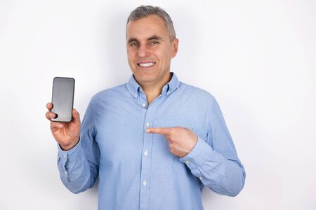 adult handsome man with grey hair wearing blue shirt demonstrates new smartphone device standing on isolated white background, modern technology conceptの写真素材