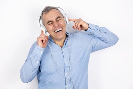 adult smiling handsome man with grey hair wearing blue shirt listens to music in headphones looks joly standing on isolated white background, positive vibes conceptの写真素材