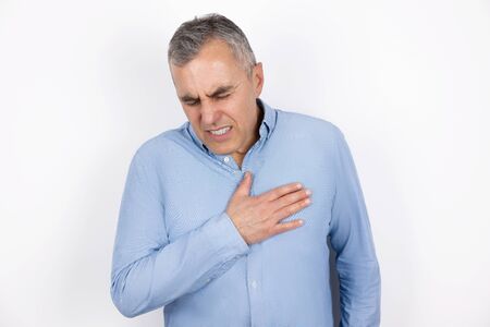 adult handsome man with grey hair wearing blue shirt touching chest with hand suffering from sudden heartache standing on isolated white, healthcare conceptの写真素材