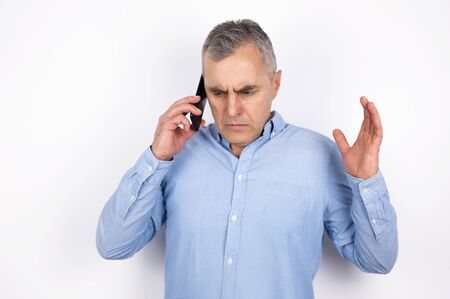 adult handsome man with grey hair wearing blue shirt having phone conversation looks desperate standing on isolated white background, modern technology conceptの写真素材
