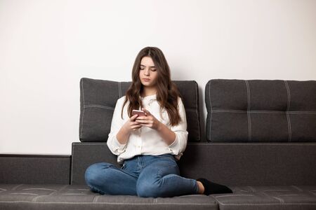 young beautiful brunette woman texting message in her smartphone sitting on the sofa in her appartment, feeling cozy at homeの写真素材