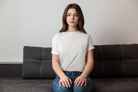 young beautiful brunette woman sitting on the sofa in her appartment holding hands on her legs looking calm, feels cozy at homeの写真素材