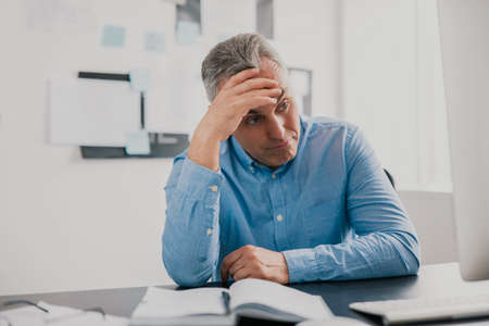 handsome tired gray-haired man sits in his office holding hand near head looking upset while working on business project , stress at workplace conceptの写真素材