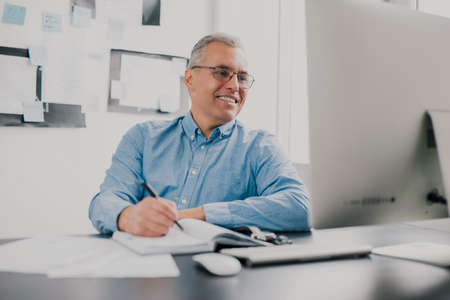 handsome gray-haired smiling man sits in his office while working on business project , checking his...
