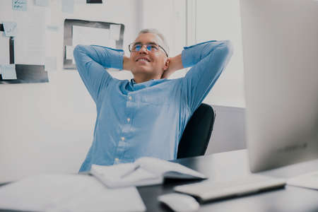 handsome gray-haired smiling man sits in his office having break while working on business project looks happy, with his hands behind head, work routine conceptの写真素材