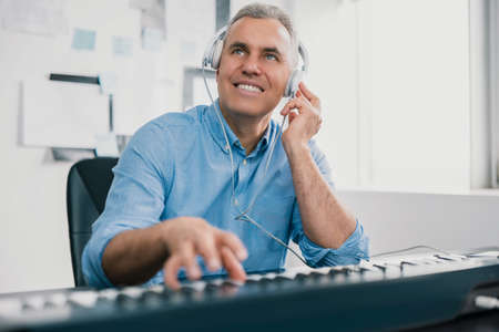 handsome gray-haired smiling man sits in his music studio playing keyboard piano wearing earphones looking excited and happy, music record concep, artの写真素材