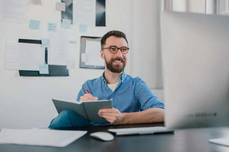 handsome young bearded man sits in his office looking happy while working on business project looks satisfied, takes notes to his agenda, work routine conceptの写真素材