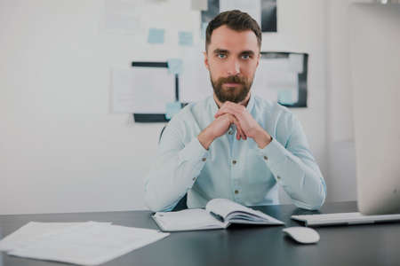 young bearded brunette man looking serious while working on business project in his modern office, holds hands near his face, work routine conceptの写真素材