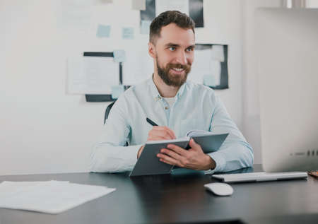 young bearded brunette smiling man looking happy while taking notes to his agenda working on business project in his modern office, work routine conceptの写真素材