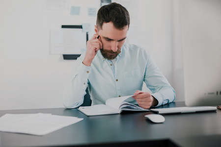 young bearded brunette man looking serious while working on business project in his modern office, holds hand with pen near his face, work routine conceptの写真素材